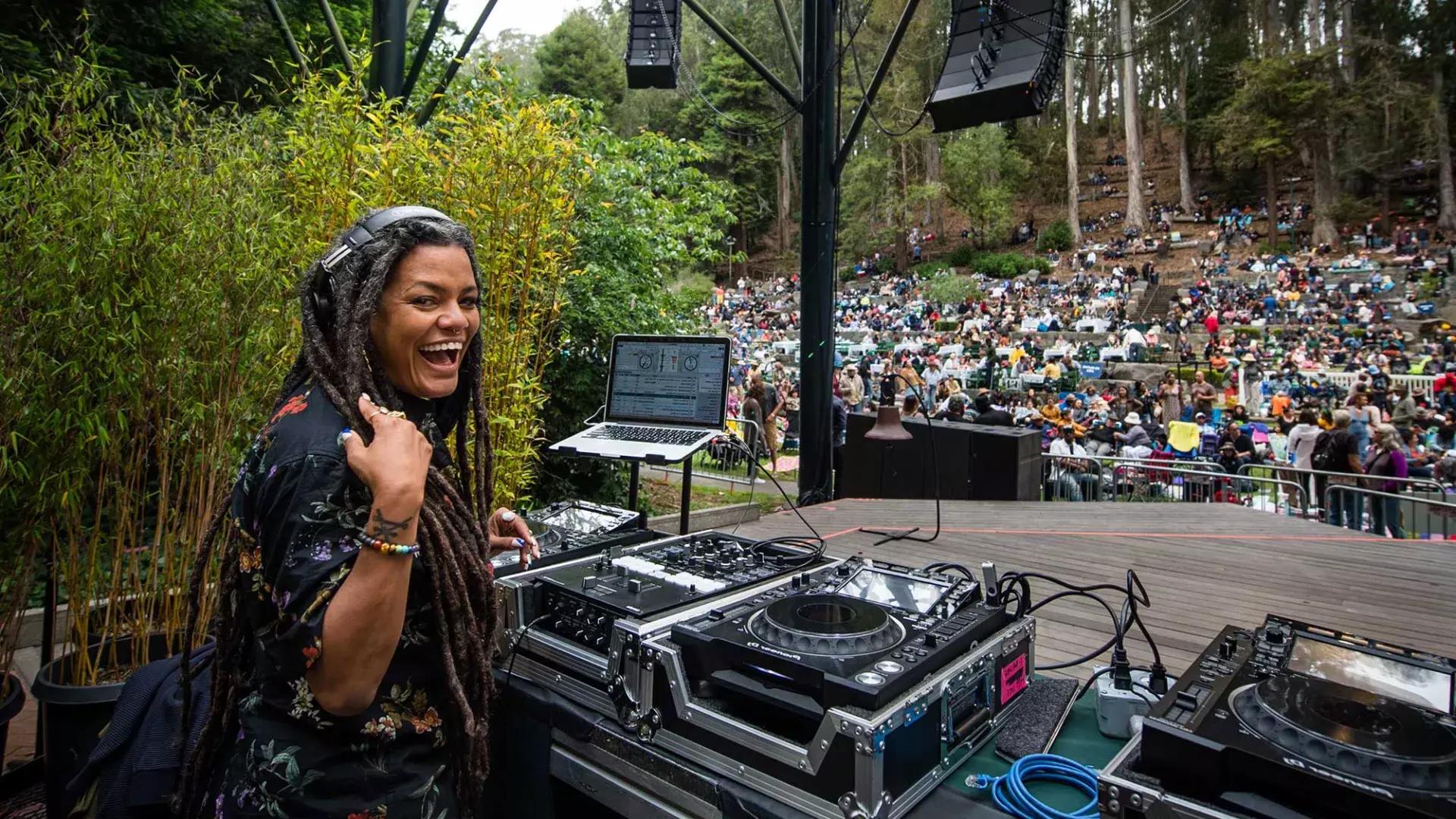 스턴 그로브 페스티벌(Stern Grove Festival)에서 DJ를 하고 있는 한 여성이 어깨 너머로 카메라를 바라보며 미소를 짓고 있습니다.