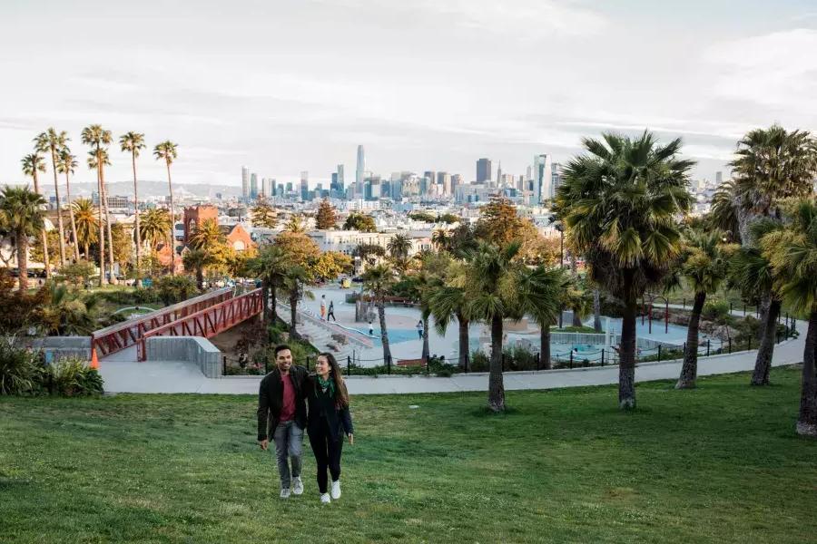 한 커플이 돌로레스 공원과 샌프란시스코 스카이라인을 뒤로하고 카메라를 향해 걸어가고 있습니다.
