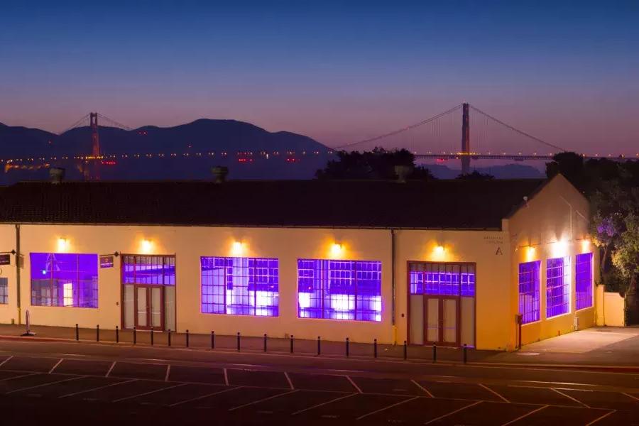 El edificio Fort Mason está iluminado con una luz interior violeta por la noche, con el puente al fondo.